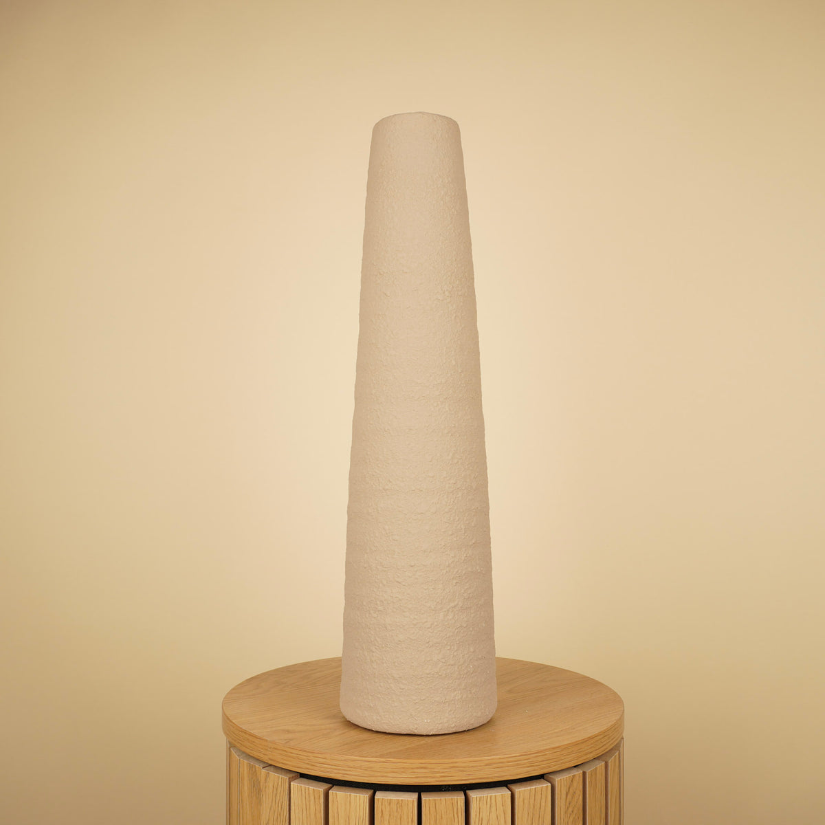 Hoge, slanke beige vaas met taps toelopende vorm en ruwe structuur op een ronde houten tafel.