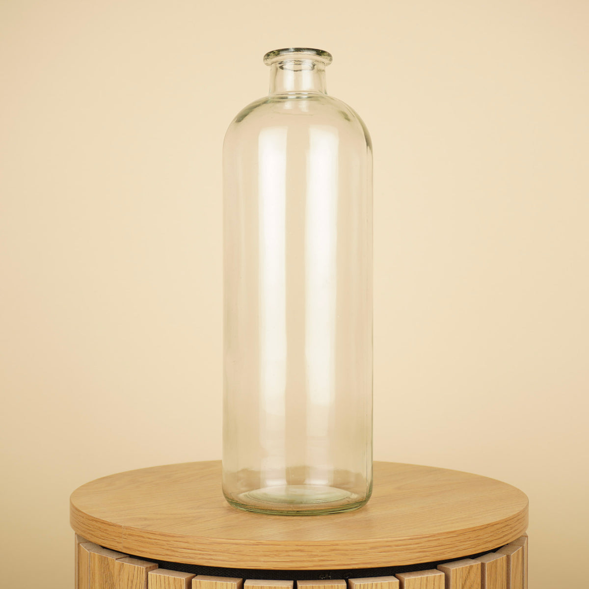 Hoge transparante glazen flesvaas met smalle hals en afgeronde schouders op een ronde houten tafel.