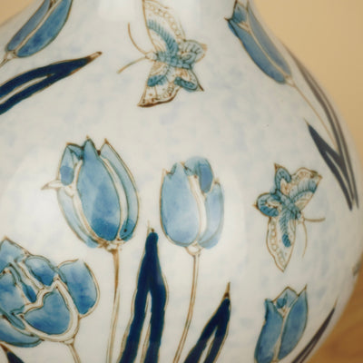 Detail van blauwe tulpen, donkerblauwe bladeren en vlinders op de glanzende witte vaas.