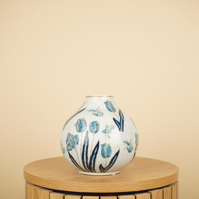 Witte vaas met ronde buik en smalle hals, versierd met blauwe tulpen en vlinders, op een houten tafel.